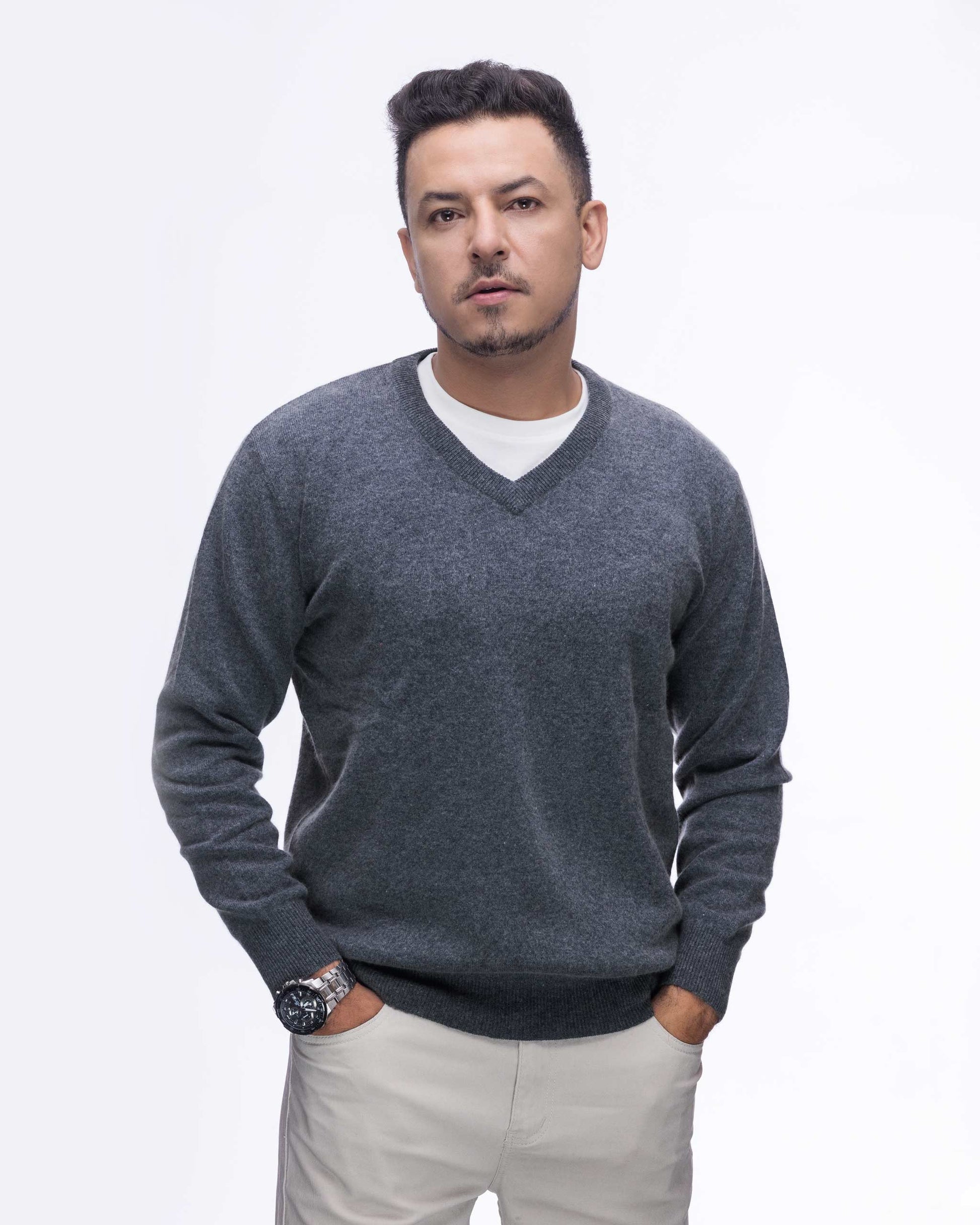 A man wearing a charcoal gray v-neck knit sweater over a white t-shirt and beige trousers, standing against a plain white background.