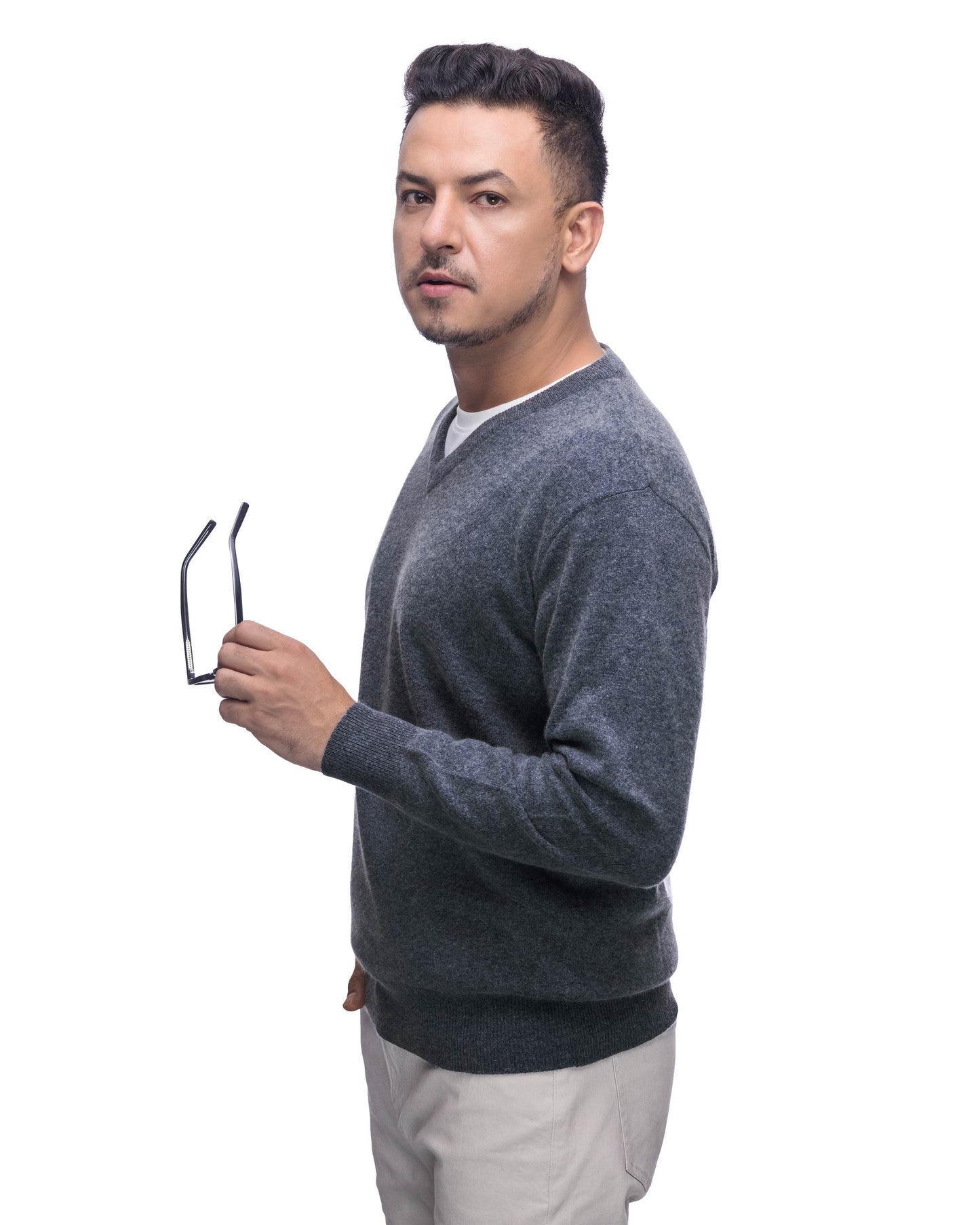 A man wearing a dark gray v-neck knit sweater and light beige trousers, holding a pair of black-rimmed glasses against a solid white background.