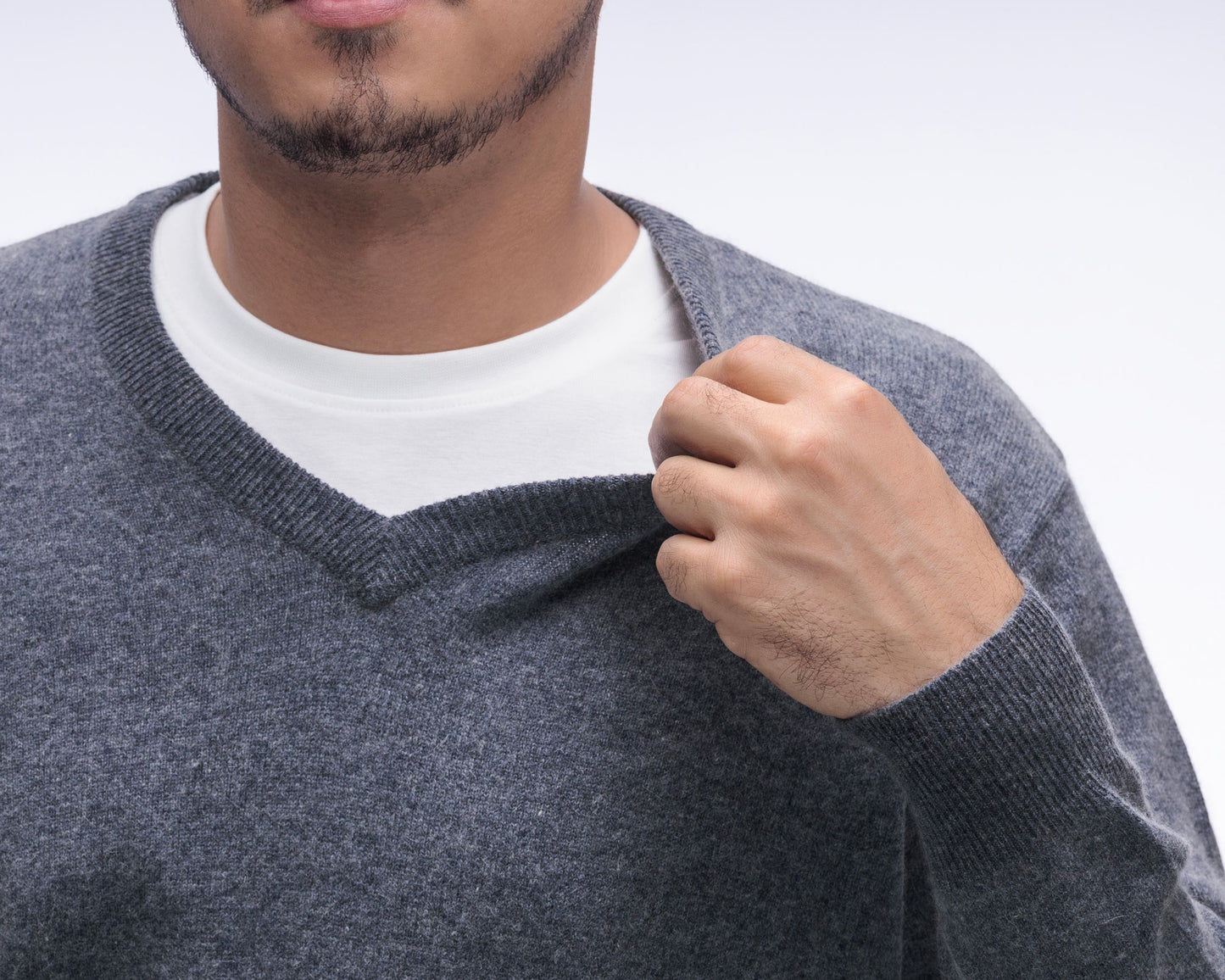 A close-up of a man wearing a heather gray v-neck sweater over a white crew-neck t-shirt, pulling the gray sweater slightly outward with his hand.