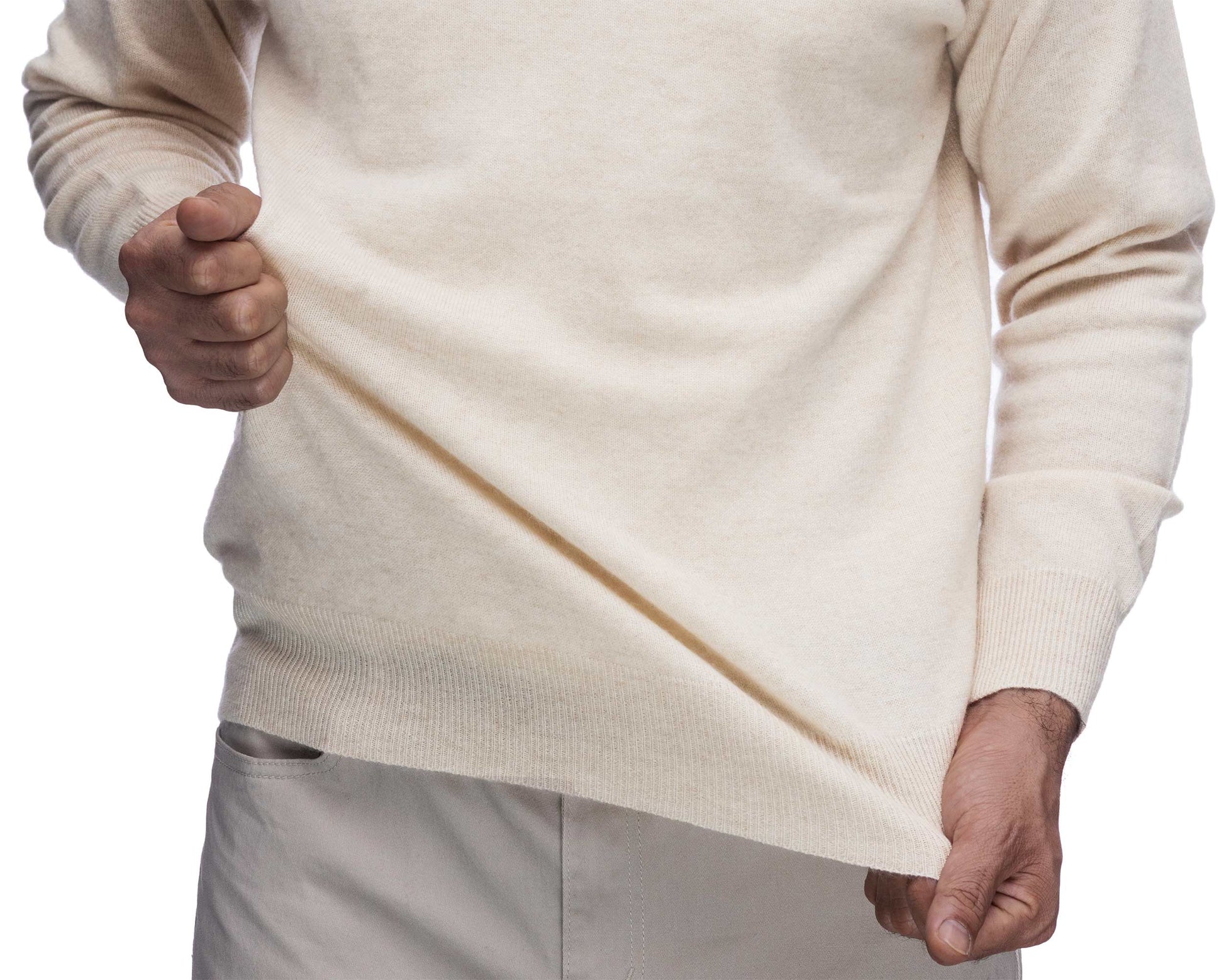 A close-up of a person wearing a cream-colored knit sweater, pulling the hem slightly downward with both hands to show the fabric's drape and texture against a white background.