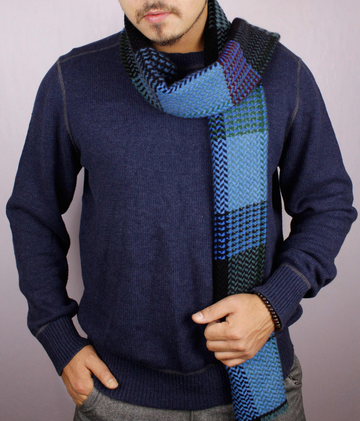 Man wearing a 100% cashmere wool herringbone scarf in shades of blue, facing the camera with one hand in his trouser pocket and the other holding the scarf draped across the front