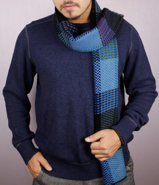 Man wearing a 100% cashmere wool herringbone scarf in shades of blue, facing the camera with one hand in his trouser pocket and the other holding the scarf draped across the front