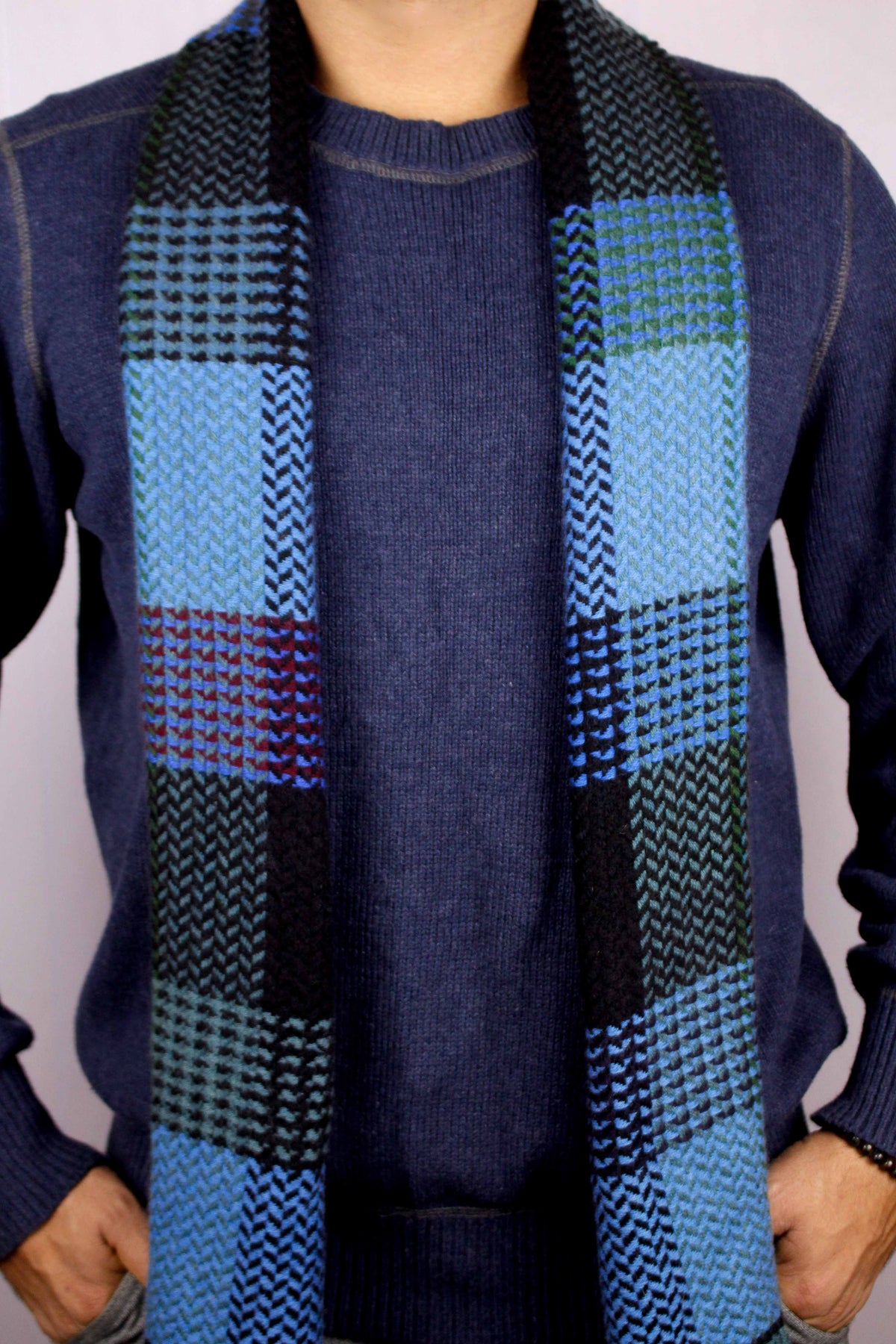 Man wearing a 100% cashmere wool herringbone scarf in shades of blue, facing the camera with the scarf draped straight down the front and both hands in his trouser pockets